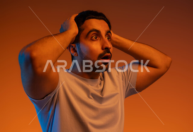 الشعور بالصدمة والذهول والانبهار، الاستغراب والتفاجؤ من شيء ما، بورتريه مقرب لشاب عربي خليجي سعودي يرتدي الزي الكاجوال و يرفع يديه على رأسه بإيماءات التعجب والدهشة، تعابير النجاح وتحقيق الأهداف, خلفية ملونة.