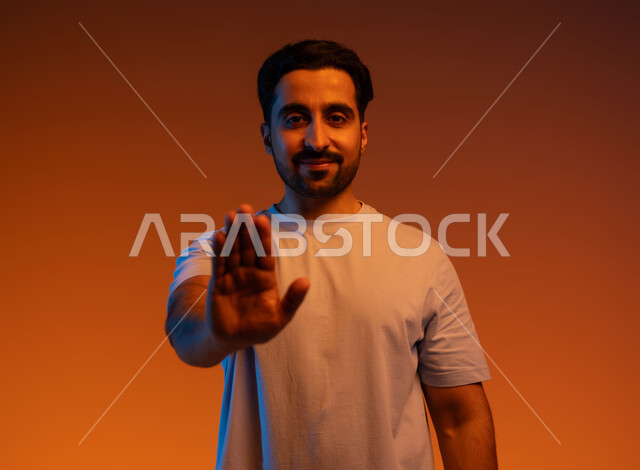 Requesting non-interference, lowering the voice and threats, gestures of non-acceptance and satisfaction, silence and stopping talking, refraining from doing a certain thing, close-up portrait of a Saudi Gulf Arab young man wearing casual clothes, raising his palm up with gestures of rejection, colored background