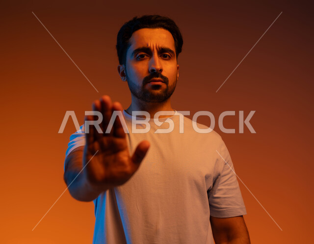 طلب عدم التدخل وخفض الصوت والتهديد, الوقوف بغضب ورفض أمر محدد, الصمت والتوقف عن الكلام, الامتناع عن فعل أمر معين, بوتريه مقرب لشاب عربي خليجي سعودي يرتدي الزي الكاجوال يرفع كفه للأعلى بإيماءات الانزعاج, خلفية ملونة
