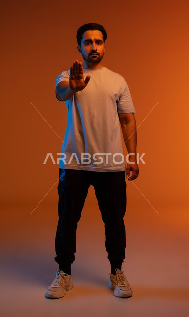 الوقوف بغضب ورفض أمر محدد, الصمت والتوقف عن الكلام, طلب عدم التدخل وخفض الصوت والتهديد, الامتناع عن فعل أمر معين, بوتريه لشاب عربي خليجي سعودي يرتدي الزي الكاجوال يرفع كفه للأعلى بإيماءات الانزعاج, صورة الجسم بالطول الكامل, خلفية ملونة