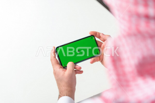 رجل أعمال سعودي خليجي يستخدام هاتفه المحمول بخلفية خضراء كروما ، تقنية و حاسب ، استخدام تقنية السمارت على الهاتف المحمول ليتحكم بالتلفاز