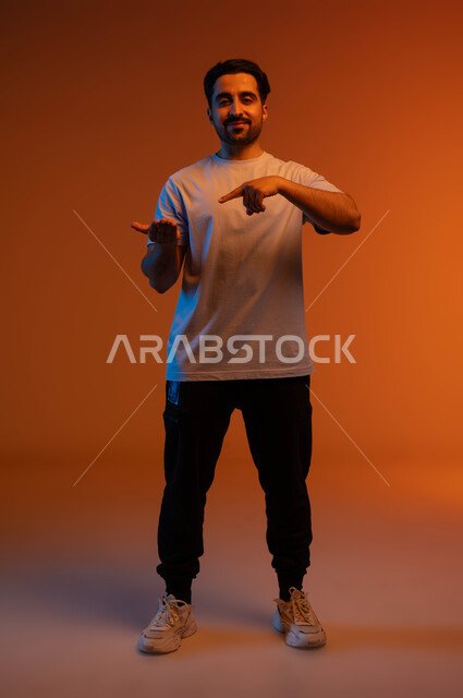 الاشارة بإصبع السبابة إلى شيء محدد, الوقوف والنظر إلى الكاميرا بإيماءات السرور, بورتريه لشاب عربي خليجي سعودي مبتسم يرتدي الزي الكاجوال يحمل بيده شيء ما, رفع وحمل عنصر معين, صورة الجسم بالطول الكامل, خلفية ملونة