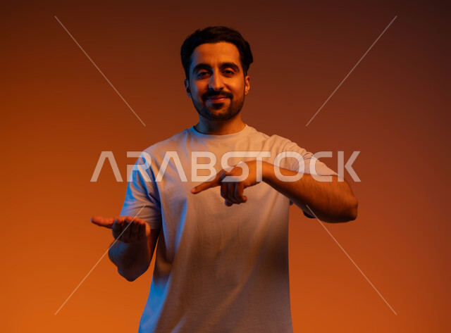 رفع وحمل عنصر معين, الوقوف والنظر إلى الكاميرا بإيماءات السرور, الاشارة بإصبع السبابة إلى شيء محدد, بورتريه مقرب لشاب عربي خليجي سعودي مبتسم يرتدي الزي الكاجوال يحمل بيده شيء ما, خلفية ملونة