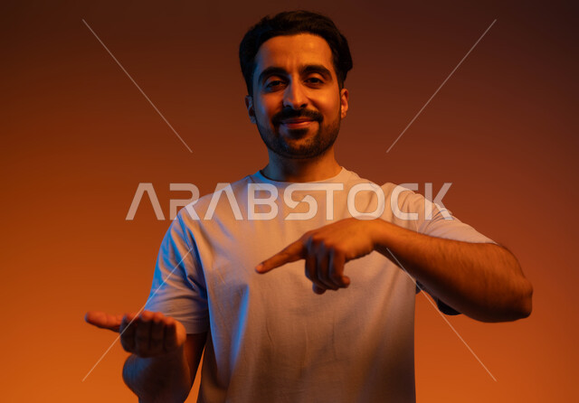 الوقوف والنظر إلى الكاميرا بإيماءات السرور, الاشارة بإصبع السبابة إلى شيء محدد, بورتريه مقرب لشاب عربي خليجي سعودي مبتسم يرتدي الزي الكاجوال يحمل بيده شيء ما, رفع وحمل عنصر معين, خلفية ملونة