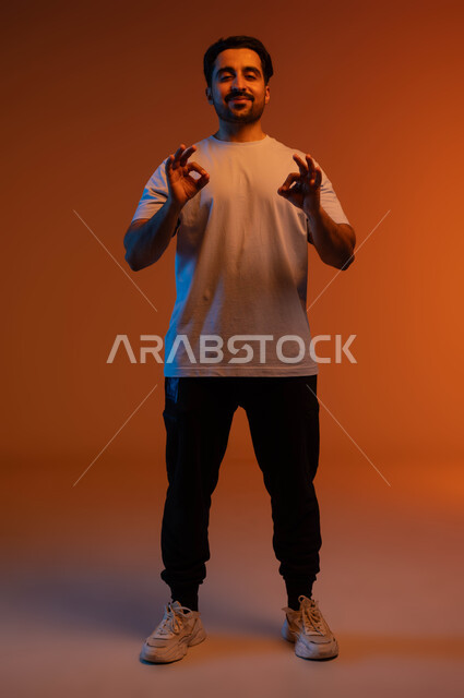 تحقيق أهداف العمل بدقة وتركيز, السرور والسعادة بإنجاز المهام, بورتريه لشاب عربي خليجي سعودي مبتسم يرتدي الزي الكاجوال ينظر إلى الكاميرا, رفع اليدين بعلامة الجودة والامتياز, صورة الجسم بالطول الكامل, خلفية ملونة