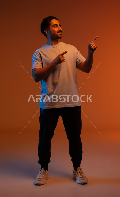 النظرة المستقبلية للأشياء, الوقوف والنظر إلى شيء ما, بورتريه لشاب عربي خليجي سعودي مبتسم يرتدي الزي الكاجوال يرفع اصبعة السبابة لجهة اليسار لتعين موقع محدد, الإشارة لمكان ما بإيماءات التخطيط, صورة الجسم بالطول الكامل, خلفية ملونة