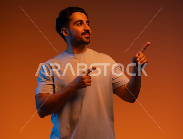 الوقوف والنظر إلى شيء ما, بورتريه مقرب لشاب عربي خليجي سعودي مبتسم يرتدي الزي الكاجوال يرفع اصبعة السبابة لجهة اليسار لتعين موقع محدد, الإشارة لمكان ما بإيماءات التخطيط, التأمل والتفكير بالأهداف المستقبلية, خلفية ملونة