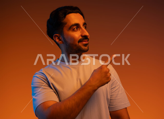 التأمل والتفكير بأمر معين, الوقوف والنظر إلى شيء ما, بورتريه مقرب لشاب عربي خليجي سعودي مبتسم يرتدي الزي الكاجوال يرفع اصبعة السبابة لجهة اليسار لتعين موقع محدد, الإشارة لمكان ما بإيماءات التخطيط, خلفية ملونة