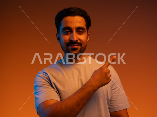 الوقوف والنظر إلى الكاميرا, بورتريه مقرب لشاب عربي خليجي سعودي يرتدي الزي الكاجوال يرفع اصبعه لتحديد وتوضيح موقع معين, الإشارة إلى هدف محدد, الذهاب لمكان ما, خلفية ملونة
