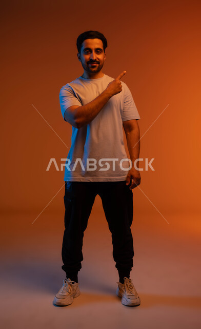 طلب الذهاب لمكان ما (إلى هناك), الإشارة إلى هدف محدد, الوقوف والنظر إلى الكاميرا, بورتريه لشاب عربي خليجي سعودي يرتدي الزي الكاجوال يرفع اصبعه لتحديد وتوضيح موقع معين, صورة الجسم بالطول الكامل, خلفية ملونة