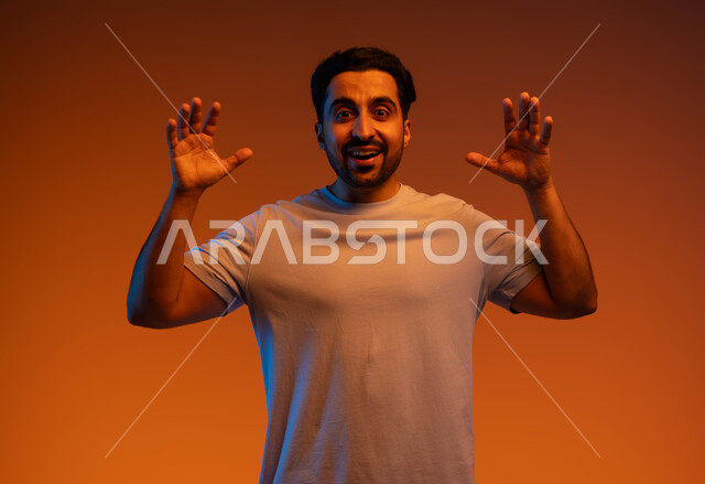 Standing with astonished gestures to see someone, joy and happiness at the completion of a specific task, pleasure in happy surprises, close-up portrait of a smiling Saudi Gulf Arab young man wearing casual clothes, raising his hands up in amazement, colorful background