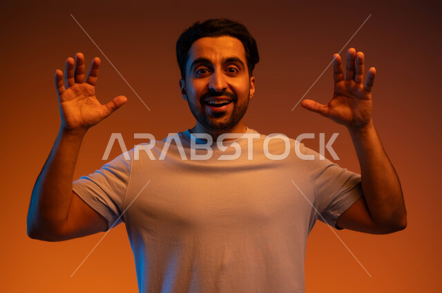 إيماءات الاستغراب لرؤية شخص ما, الفرح والسعادة بانتهاء إنجاز عمل محدد, السرور بالمفاجآت السعيدة, بورتريه مقرب لشاب عربي خليجي سعودي مبتسم يرتدي الزي الكاجوال يرفع يديه للأعلى بدهشة, خلفية ملونة