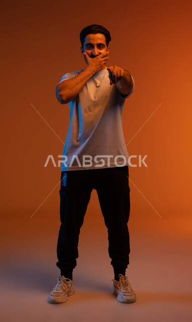 المفاجأة لرؤية شخص ما, الدهشة والذهول والاستغراب وتحديد الشيء المقابل, الوقوف باستقامة والنظر إلى الكاميرا, بورتريه لشاب عربي خليجي سعودي يرتدي الزي الكاجوال يضع يده على فمه ويشير بإصبعه السبابة لمكان معين, صورة الجسم بالطول الكامل, خلفية ملونة