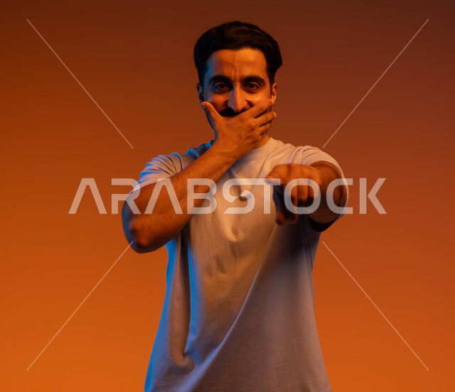 المفاجأة لرؤية شخص ما, الدهشة والذهول والاستغراب وتعين مكان محدد, الوقوف باستقامة والنظر إلى الكاميرا, بورتريه لشاب عربي خليجي سعودي يرتدي الزي الكاجوال يضع يده على فمه ويشير بإصبعه السبابة لتحديد الشيء المقابل, خلفية ملونة