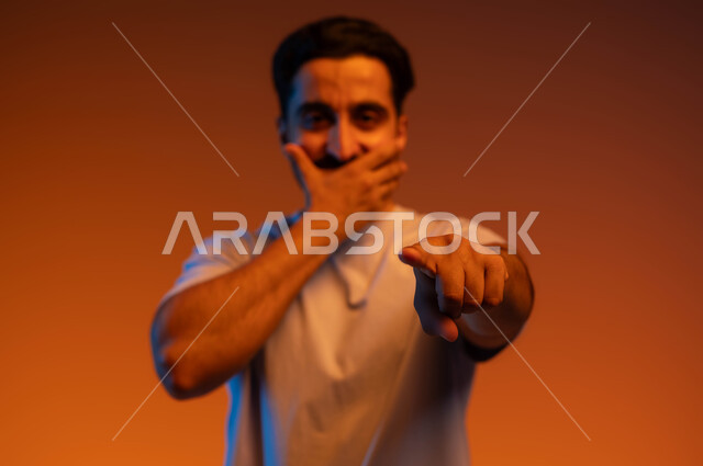 الدهشة والذهول والاستغراب وتعين مكان محدد, المفاجأة لرؤية شخص ما, الوقوف باستقامة والنظر إلى الكاميرا, بورتريه لشاب عربي خليجي سعودي يرتدي الزي الكاجوال يضع يده على فمه ويشير بإصبعه السبابة لتحديد الشيء المقابل, خلفية ملونة