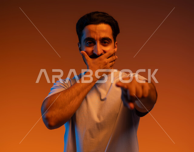 Astonishment and astonishment, specifying a specific place, surprise to see someone, standing up straight and looking at the camera, portrait of a Saudi Gulf Arab young man wearing a casual outfit, placing his hand over his mouth and pointing with his index finger to identify the corresponding object, colored background