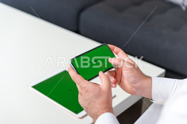 رجل أعمال سعودي خليجي يستخدام هاتفه المحمول بخلفية خضراء كروما ، تقنية و حاسب ، استخدام تقنية السمارت على الهاتف المحمول ليتحكم بالتلفاز