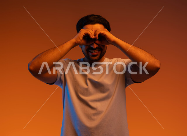 البشاشة والسرور برؤية شيء ما, محاولة تقريب الأشياء البعيدة, التخطيط للأهداف والطموحات المستقبلية, بورتريه مقرب لشاب عربي خليجي سعودي مبتسم يرتدي الزي الكاجوال يضع يديه على عينيه, حب الاكتشاف والمعرفة والمغامرة, خلفية ملونة