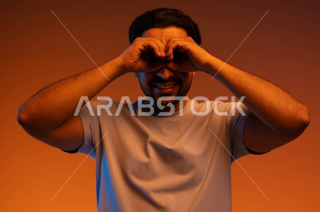 محاولة رؤية الأشياء عن قرب, الوقوف بسرور وبشاشة, التخطيط للأهداف والطموحات المستقبلية, بورتريه لشاب عربي خليجي سعودي مبتسم يرتدي الزي الكاجوال يضع يديه على عينيه, حب الاكتشاف والمعرفة والمغامرة, خلفية ملونة