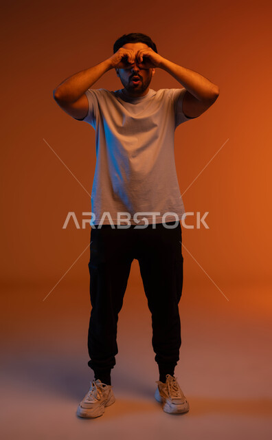 المعرفة والاكتشاف والمغامرة, رؤية الأشياء عن قرب, التخطيط والرؤية المستقبلية للأهداف والطموحات, بورتريه لشاب عربي خليجي سعودي مبتسم يرتدي الزي الكاجوال يضع يديه على عينه على شكل عدسة, صورة الجسم بالطول الكامل, خلفية ملونة