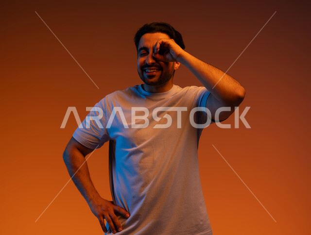 رؤية الأشياء عن قرب, التخطيط والرؤية المستقبلية للأهداف والطموحات, بورتريه مقرب لشاب عربي خليجي سعودي مبتسم يرتدي الزي الكاجوال يضع يديه على عينه على شكل عدسة, حب المعرفة والاكتشاف والمغامرة, خلفية ملونة