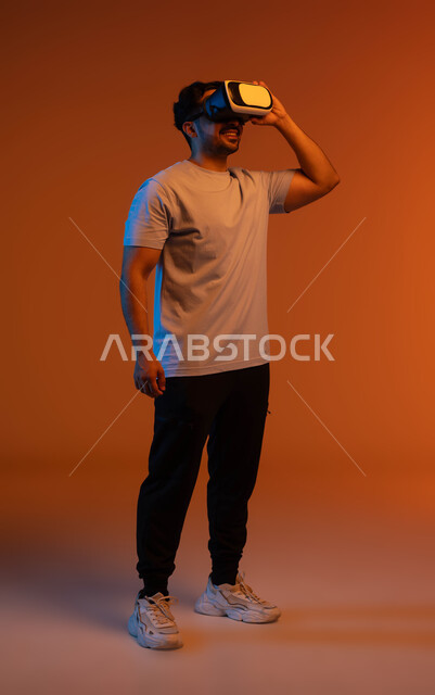 التفاعل والاندماج ومحاكاة العالم الخيالي, بورتريه لشاب عربي خليجي سعودي مبتسم يرتدي الزي الكاجوال يمسك بيده نظارة الواقع الافتراضي, التسلية ومشاهدة الأفلام في وقت الفراغ, استخدام التقنيات والأجهزة المتطورة, صورة الجسم بالطول الكامل, خلفية ملونة