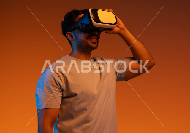 التسلية والاستمتاع بمشاهدة الأفلام و محاكاة العالم الخيالي، استخدام التقنيات والاجهزة الحديثة, بورتريه مقرب لشاب عربي سعودي خليجي مبتسم يرتدي الزي الكاجوال ونظارة الواقع الافتراضي ثلاثية الأبعاد بإيماءات السرور، مفهوم التفاعل و الاندماج، خلفية ملونة .