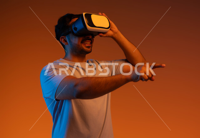 الإشارة إلى شيء ما بأصبع السبابة، الاندماج والتفاعل و محاكاة العالم الخيالي، بورتريه مقرب لشاب عربي سعودي خليجي يرتدي الزي الكاجوال ونظارة في آر الافتراضية ثلاثية الأبعاد يحاول ملامسة العناصر الهولوجرامية بإيماءات النقر، خلفية ملونة .