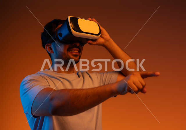 استخدام حاسة اللمس لملامسة عناصر هولوجرامية بإيماءات النقر, الاندماج والتفاعل و محاكاة العالم الخيالي، بورتريه مقرب لشاب عربي سعودي خليجي يرتدي الزي الكاجوال ونظارة في آر الافتراضية ثلاثية الأبعاد يشير إلى شيء ما بأصبعه السبابة، خلفية ملونة .