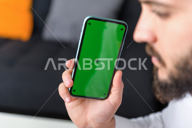 رجل أعمال سعودي خليجي يستخدام هاتفه المحمول بخلفية خضراء كروما ، تقنية و حاسب ، استخدام تقنية السمارت على الهاتف المحمول ليتحكم بالتلفاز