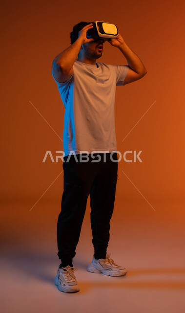 Integrating and interacting with virtual reality games, watching movies and simulating the imaginary world, gestures of astonishment and astonishment, a close-up portrait of a young Saudi Arabian Gulf man wearing casual clothes and virtual 3D VR glasses, full-length body image, colored background