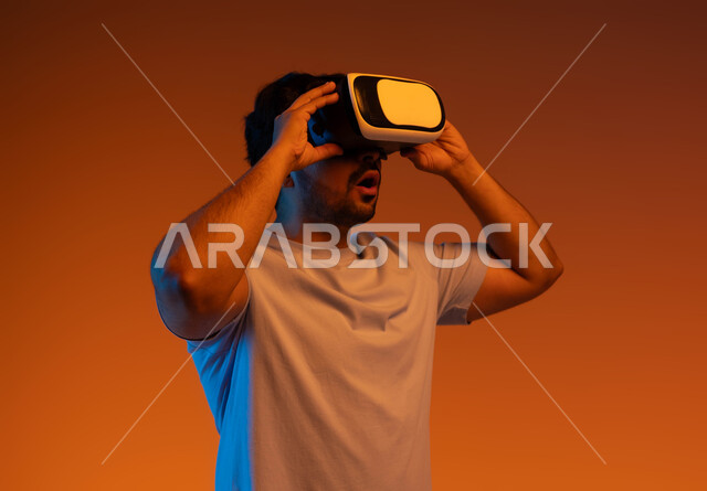 Interacting and merging with virtual reality games, watching movies and simulating the imaginary world, gestures of astonishment and astonishment, using modern technologies and devices, a close-up portrait of a young Saudi Arabian Gulf man wearing casual clothes and 3D virtual VR glasses, a colorful background