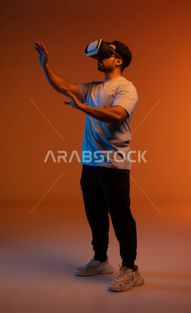التفاعل و الاندماج مع ألعاب الواقع الافتراضي، مفهوم التطور التقني والتكنولوجي, محاكاة العالم الخيالي، بورتريه مقرب لشاب عربي سعودي خليجي يتدي الزي الكاجوال ونظارة في آر الافتراضية ثلاثية الأبعاد يحاول لمس شيء ما، صورة الجسم بالطول الكامل، خلفية ملونة .
