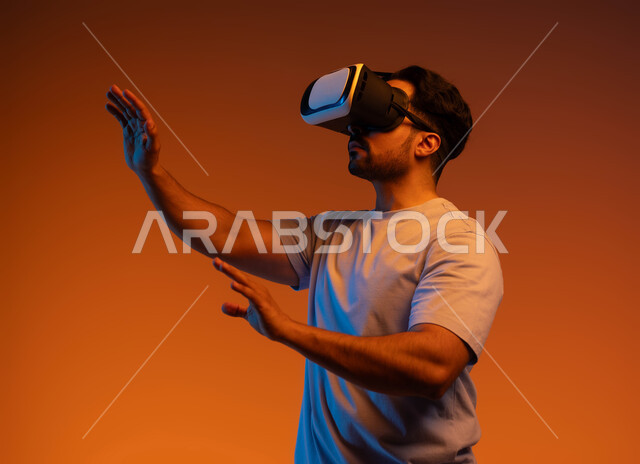 مفهوم التطور التقني والتكنولوجي, محاكاة العالم الخيالي والمتعة بمشاهدة الأفلام، إيماءات التفاعل و الاندماج، بورتريه مقرب لشاب عربي سعودي خليجي يتدي الزي الكاجوال ونظارة الواقع الافتراضي ثلاثية الأبعاد يحاول لمس شيء ما، خلفية ملونة .