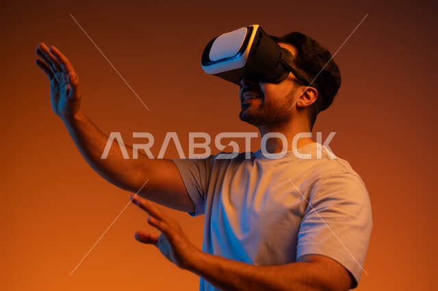 التفاعل و الاندماج  ومحاكاة العالم الخيالي، الاستمتاع بمشاهدة الأفلام، مفهوم التطور التقني التكنولوجي, بورتريه مقرب لشاب عربي سعودي خليجي يتدي الزي الكاجوال ونظارة الواقع الافتراضي ثلاثية الأبعاد يحاول لمس شيء ما، خلفية ملونة .