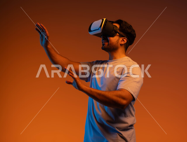 Simulating the imaginary world and the pleasure of watching movies, gestures of interaction and integration, the concept of technological development, a close-up portrait of a young Saudi Arabian Gulf man wearing a casual outfit and 3D virtual reality glasses trying to touch something, a colorful background