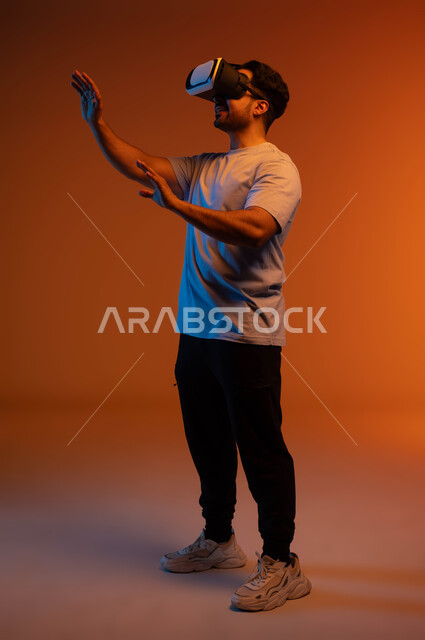 Enjoying watching movies and simulating the imaginary world, gestures of interaction and integration, the concept of technological development, a portrait of a young Saudi Arabian Gulf man wearing a casual outfit and 3D virtual reality glasses trying to touch something, a full-length body image, a colored background
