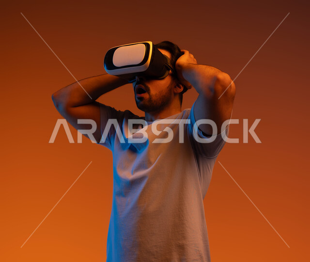 إيماءات الدهشة والذهول، التطور التقني والتكنولوجي، مفهوم التفاعل و الاندماج، بورتريه لشاب عربي سعودي خليجي يتدي الزي الكاجوال ونظارة الواقع الافتراضي ثلاثية الأبعاد يضع يديه على رأسه، المتعة بمشاهدة الأفلام و محاكاة العالم الخيالي، خلفية ملونة .