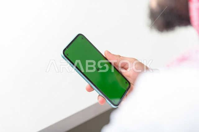 رجل أعمال سعودي خليجي يستخدام هاتفه المحمول بخلفية خضراء كروما ، تقنية و حاسب ، استخدام تقنية السمارت على الهاتف المحمول ليتحكم بالتلفاز
