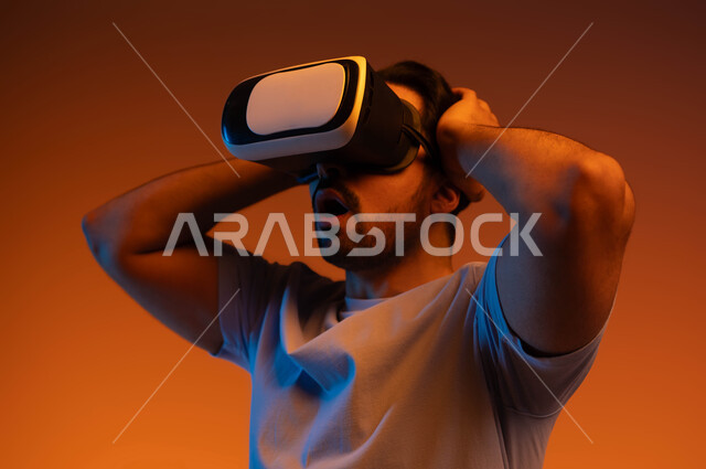 إيماءات الذهول والمفاجأة، التطور التقني والتكنولوجي، مفهوم التفاعل و الاندماج، بورتريه لشاب عربي سعودي خليجي يتدي الزي الكاجوال ونظارة الواقع الافتراضي ثلاثية الأبعاد يضع يديه على رأسه، المتعة بمشاهدة الأفلام و محاكاة العالم الخيالي، خلفية ملونة .