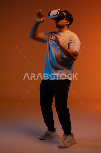 Technical and technological development, the concept of interaction and integration, a portrait of a young Saudi Arab Gulf man wearing casual clothes and 3D virtual reality glasses trying to touch something, having fun watching movies and simulating the imaginary world, full-length body image, colored background