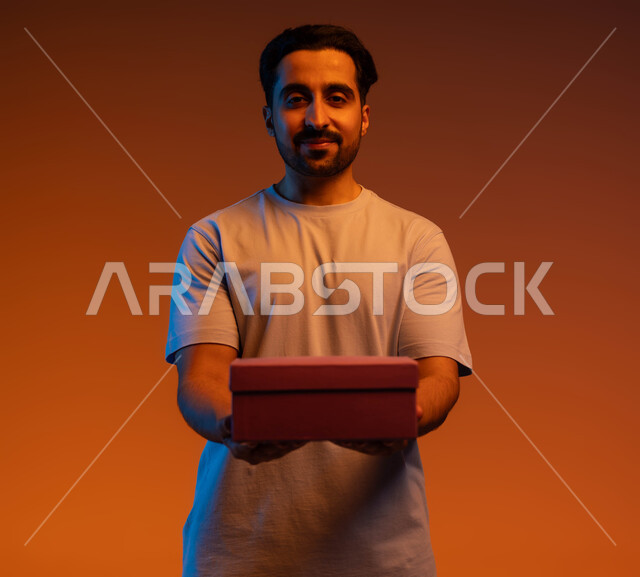 السعادة بتقديم الهدايا والتهنئة, أعياد ومناسبات ومفاجآت سعيدة, مفهوم الإهداء والتقدير, بورتريه مقرب لشاب عربي سعودي خليجي يرتدي الزي الكاجوال يمسك بيده علبة مغلقة, خلفية ملونة