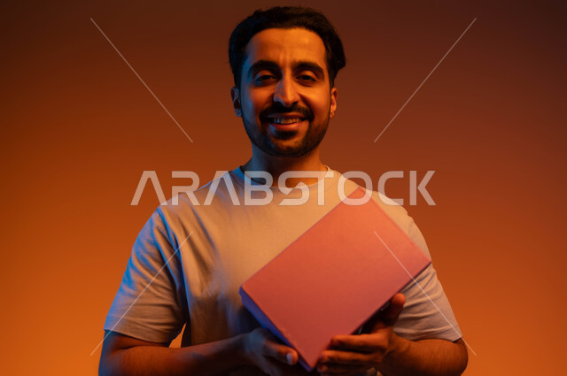 الشعور بالامتنان بالحصول على الهدية, مفهوم الإهداء والتقدير, أعياد ومناسبات ومفاجآت سعيدة, بورتريه مقرب لشاب عربي سعودي خليجي مبتسم  يرتدي الزي الكاجوال يمسك بيده علبة مغلقة .خلفية ملونة