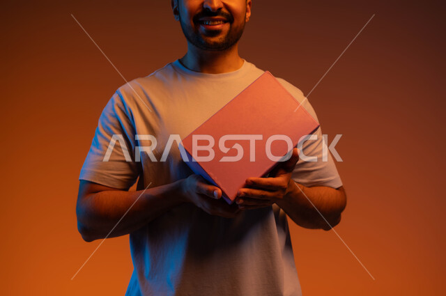 Surprises, holidays and happy occasions, the concept of gifting and appreciation, acquiring and choosing gifts, gestures of pleasure in receiving special gifts, a close-up portrait of a smiling Saudi Arab Gulf man wearing a casual outfit holding a closed box in his hands, a colored background