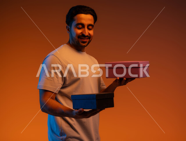 مناسبات وأعياد سعيدة, إيماءات السرور بالحصول على الهدايا تذكارية, مفهوم الإهداء والتقدير, بورتريه مقرب لشاب عربي سعودي خليجي يرتدي الزي الكاجوال يحمل بيديه علب مغلقة, اقتناء واختيار الهدايا, خلفية ملونة