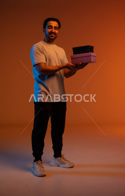 الوقوف بإيماءات السرور والنظر الى الكاميرا, أعياد ومفاجآت سعيدة, اقتناء وتقديم الهدايا بالمناسبات العائلية, بورتريه لشاب عربي سعودي خليجي يرتدي الزي الكاجوال يحمل بيده علب هدايا مغلقة, صورة الجسم بالطول الكامل, خلفية ملونة