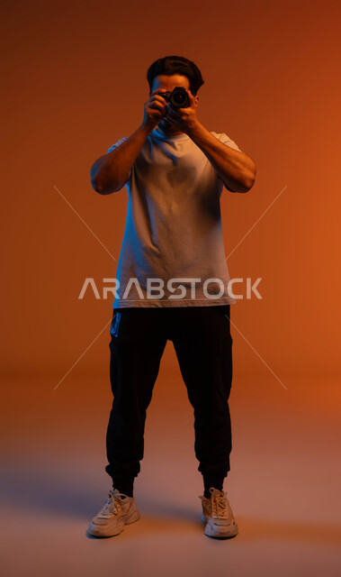 Using high-resolution digital cameras, practicing the hobby of photography, a portrait of a Saudi Gulf Arab young man wearing casual clothes holding a photographic camera in his hand, a professional photographer taking souvenir photos, full-length body portrait, colored background