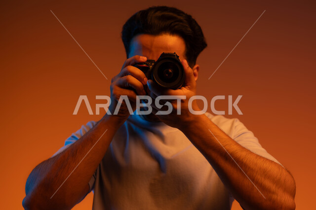 ممارسة هواية التصوير, بورتريه مقرب لشاب عربي خليجي سعودي يرتدي الزي الكاجوال يمسك بيده كاميرا فوتوغرافية, استخدام كاميرات رقمية عالية الدقة, مصور محترف يقوم بالتقاط صور تذكارية, خلفية ملونة