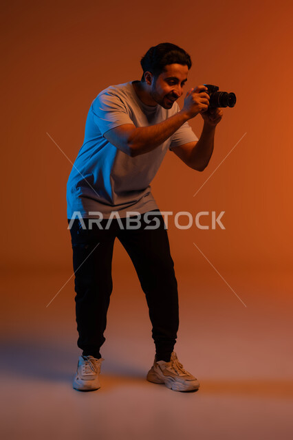 ممارسة هواية التصوير, مصور محترف يقوم بالتقاط صور تذكارية، وظائف ومهن سعودية, استخدام كاميرات رقمية عالية الدقة, بورتريه من الجانب لرجل عربي خليجي سعودي يرتدي الزي الكاجوال يقوم بالتقاط صور عبر كاميرا فوتوجرافية, صورة الجسم بالطول الكامل, خلفية ملونة