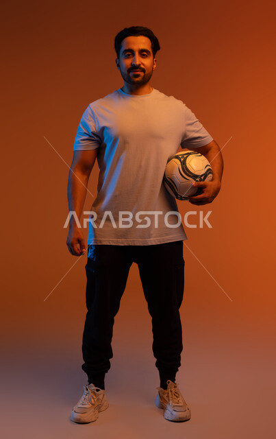 الوقوف باستقامة و النظر إلى الكاميرا، الاستعداد لبدء المباراة، ممارسة الهواية المفضلة، بورتريه للاعب عربي سعودي خليجي يرتدي الزي الكاجوال يحمل كرته بيده، احتراف الرياضات الجماعية، احتراف اللعب بالكره القدم، التسلية و الترفيه بالرياضات الحركية، صورة الجسم بالكامل، خلفية ملونة